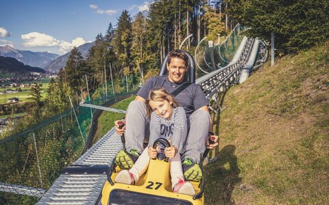 Ein Mann und ein Mädchen fahren mit einer Sommerrodelbahn.