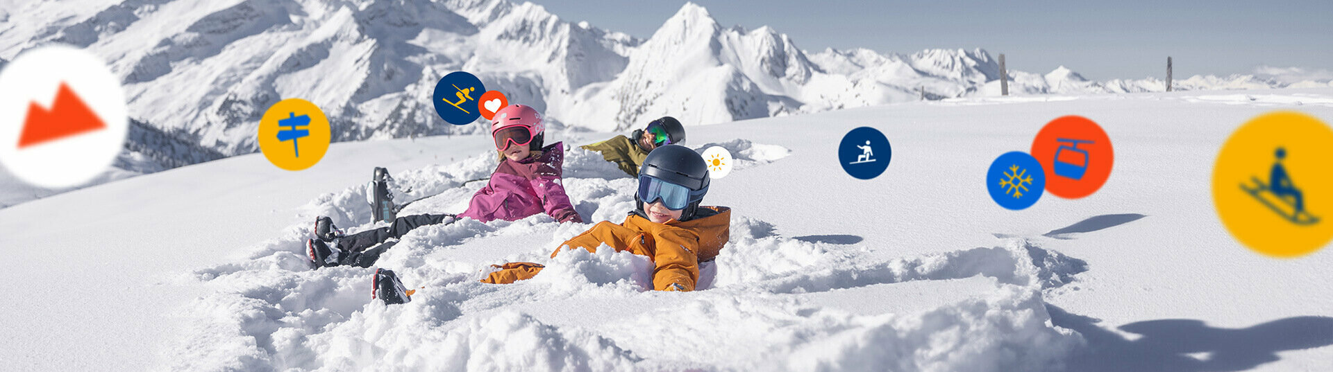 Ein Mann und zwei Kinder in Skibekleidung liegen im Schnee, im Hintergrund eine verschneite Alpenlandschaft.