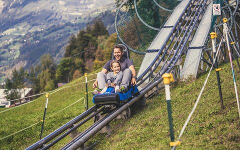 Ein Mann und ein Mädchen fahren mit einer Sommerrodelbahn.