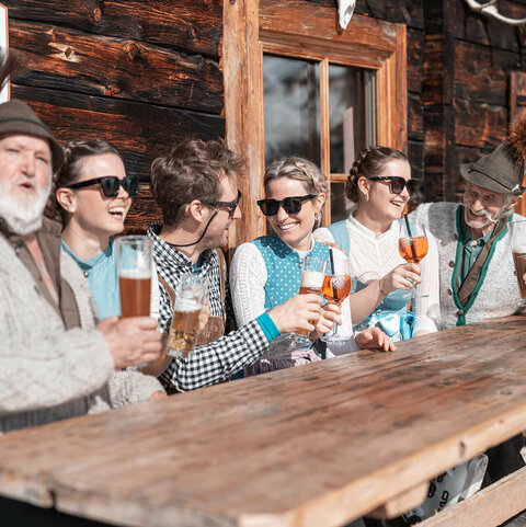 Drei Männer und drei Frauen in Trachtenkleidung trinken Bier und Aperol bei einer Hütte