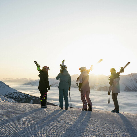 Vier Leute mit den Skiern auf den Schultern schauen in den Sonnenaufgang