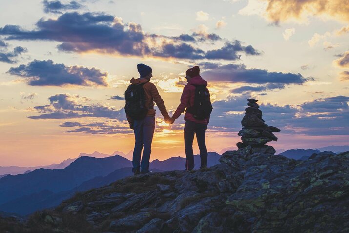 Ein Mann und eine Frau stehen händchenhaltend am kreuzjoch und beobachten einen Sonnenaufgang