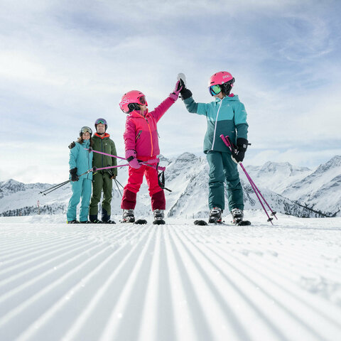 Eine Familie auf der Skipiste die Kinder machen High Five die Eltern stehen dahinter Arm in Arm