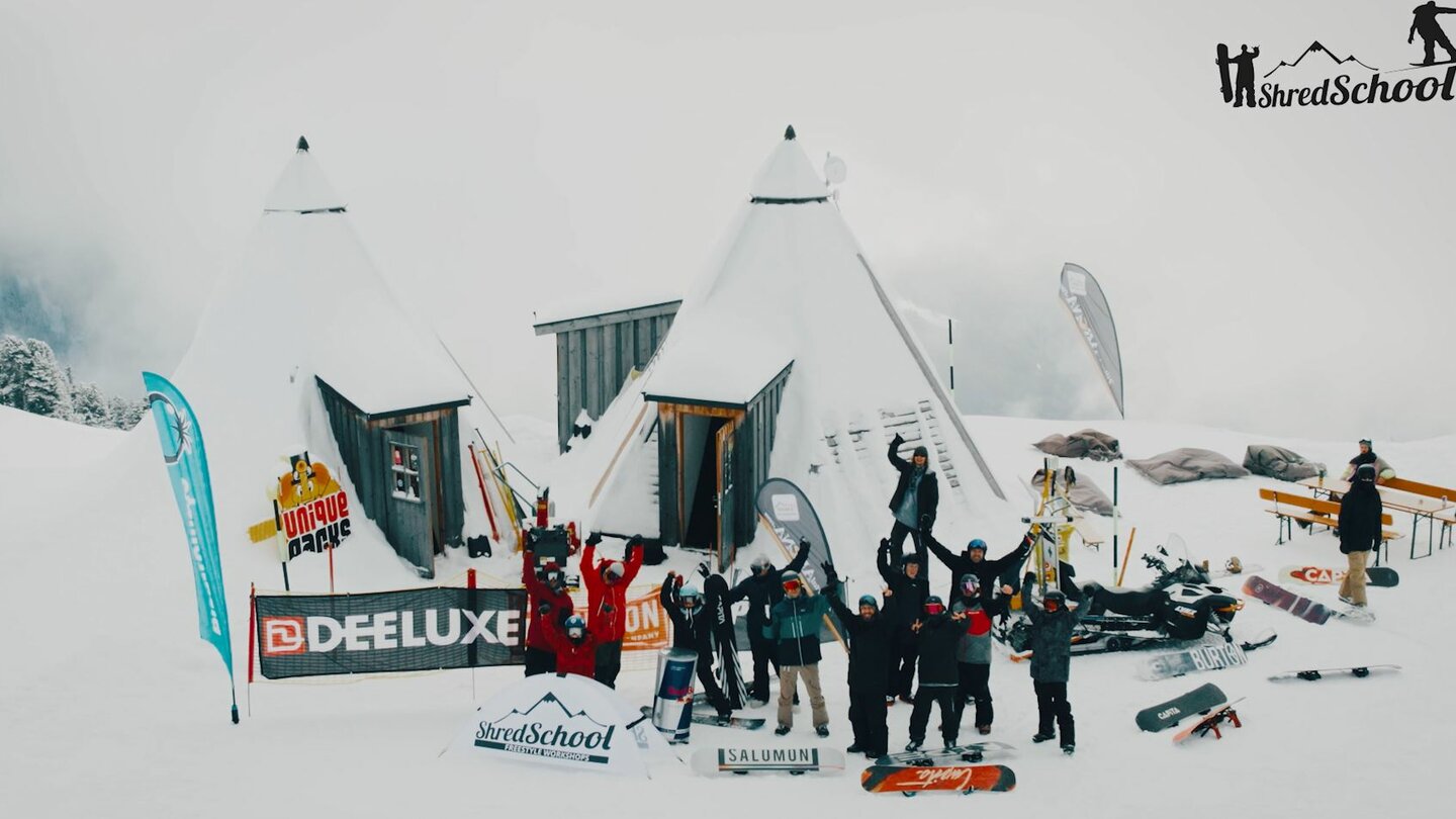 Gruppe von Wintersportlern vor zwei Tipi-Zelten im Schnee mit Snowboards, umgeben von winterlicher Berglandschaft.
