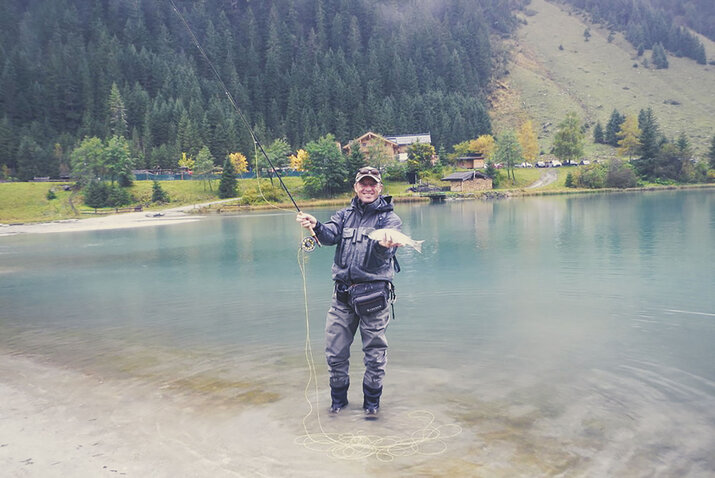 Ein Angler steht an einem See und hält einen Fisch in der Hand.