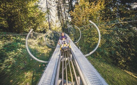 Ein Mann und ein Mädchen fahren mit einer Sommerrodelbahn.
