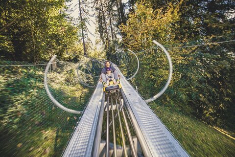 Ein Mann und ein Mädchen fahren mit einer Sommerrodelbahn.