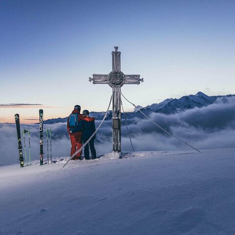 Zwei Skifahrer stehen neben dem Gipfelkreuz und genießen die Aussicht