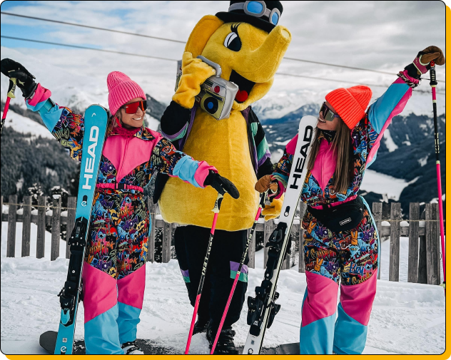 Zwei Frauen mit Skiern und bunten Skianzügen stehen neben dem Maskottchen Funty. 